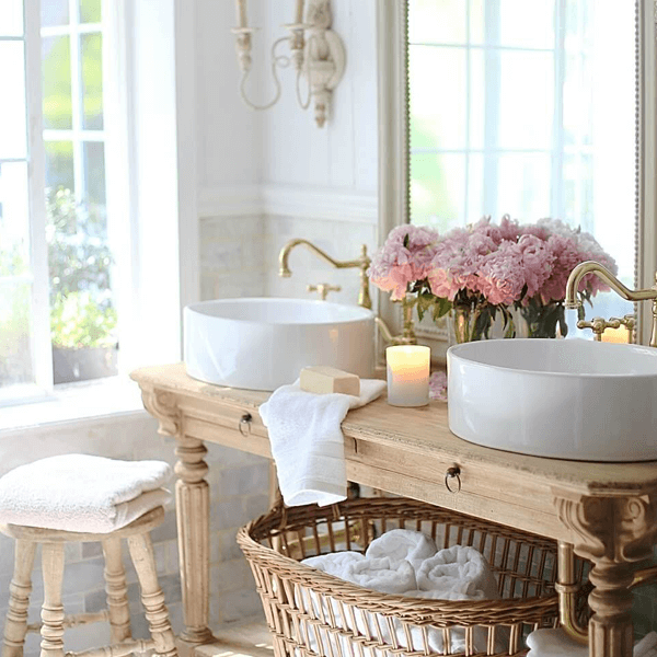 A French Provincial styled bathroom with two white basins sitting on a rustic table and wicker basket below